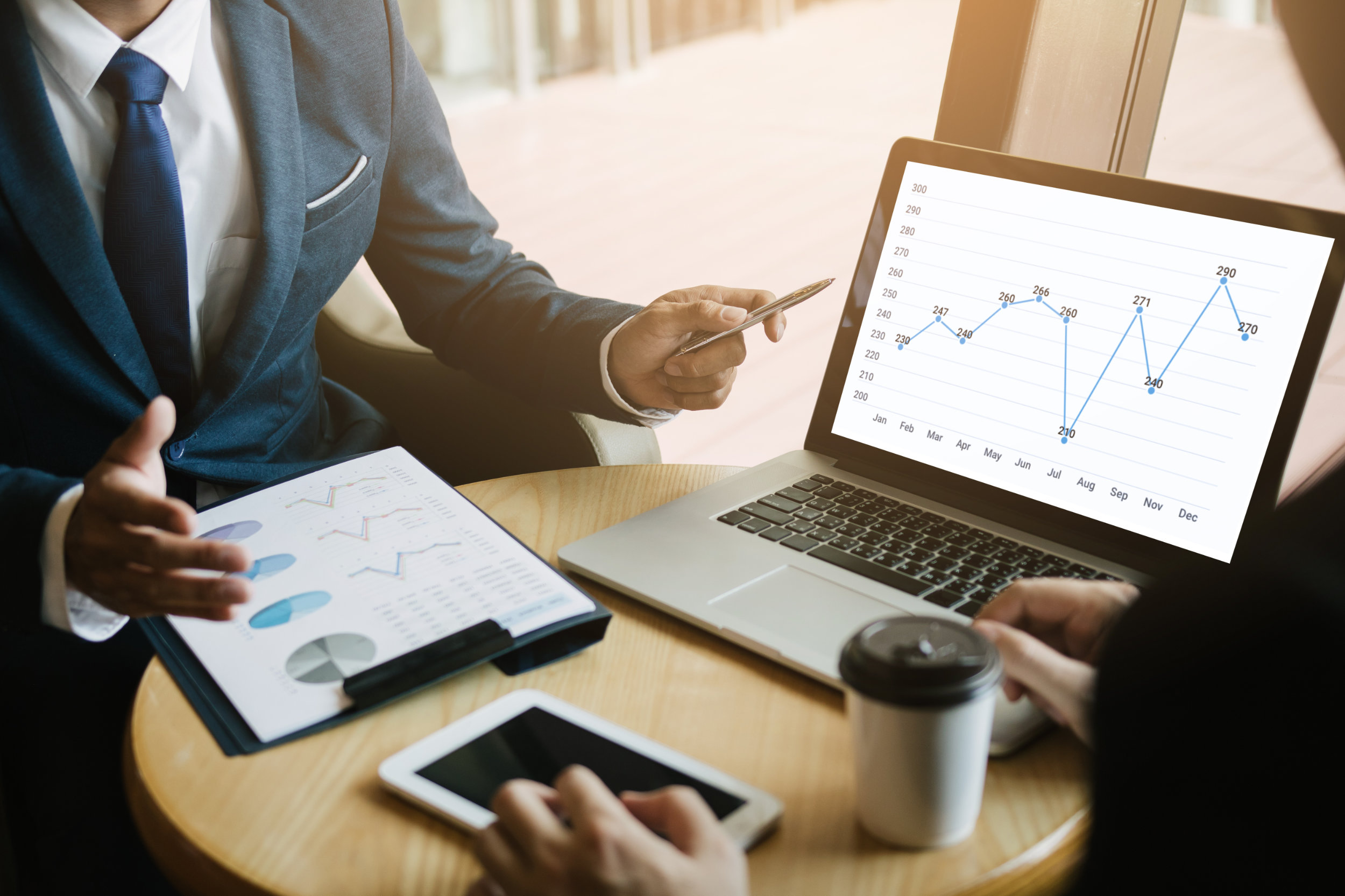 Business professionals discussing charts and graphs displayed on a laptop and a clipboard, with a coffee cup and tablet on the table.